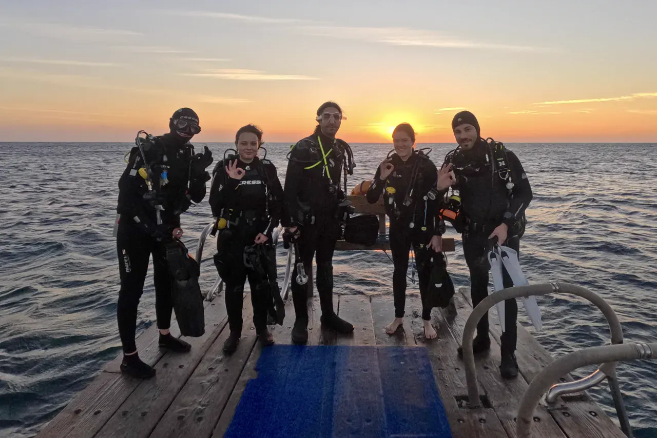 Sechs Taucherinnen und Taucher in voller Ausrüstung stehen bei Sonnenuntergang auf einem Steg am Meer